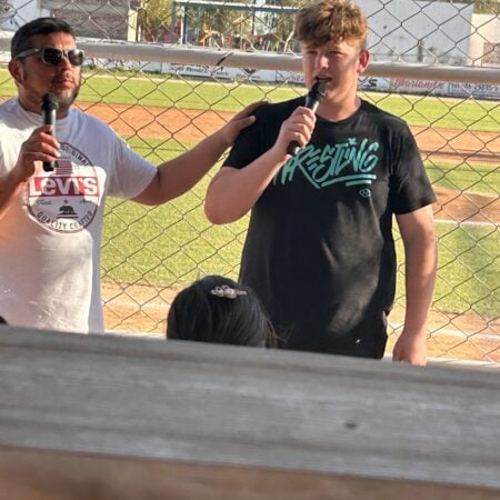 Two men speaking into microphones near a baseball field fence