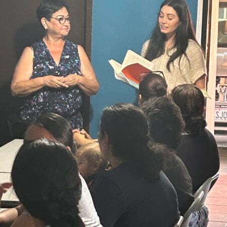 A young woman holding an open book and speaking to a gathered group of women