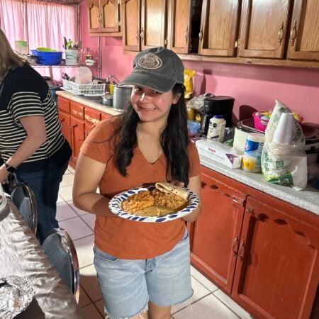 A smiling young woman in a cap holding a paper plate with a hot meal in a kitchen