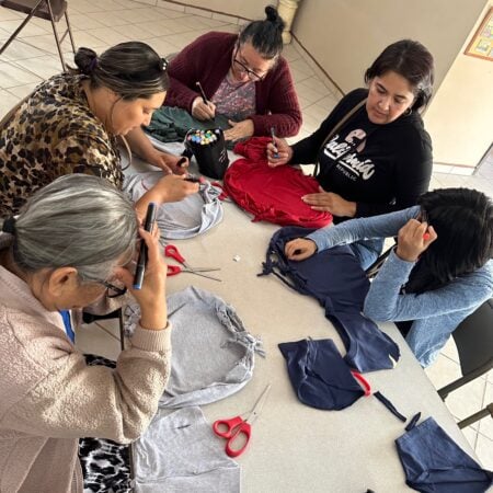 Group of women sitting around a table cutting gray and dark fabrics with scissors