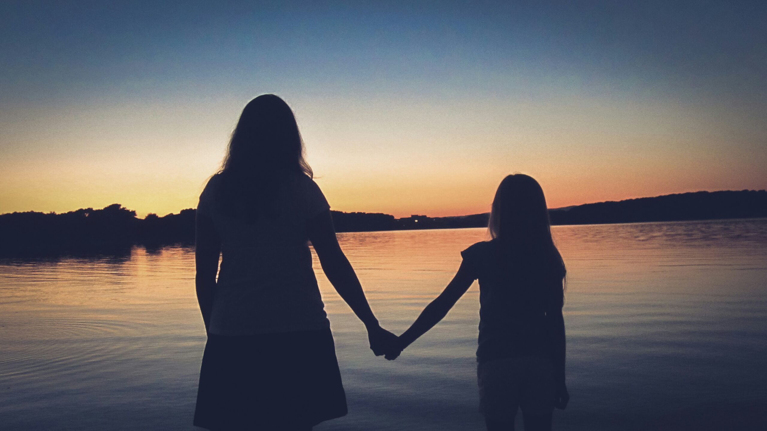 Silhouette of a mother and daughter holding hands by a lake at sunset